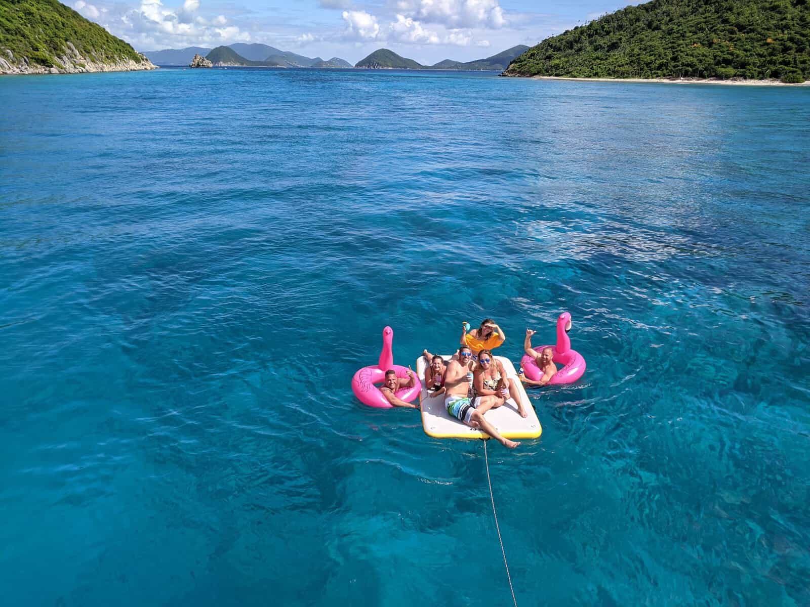 Group of friends floating on a mat and inflatable flamingos in teal blue water between green islands in the Caribbean