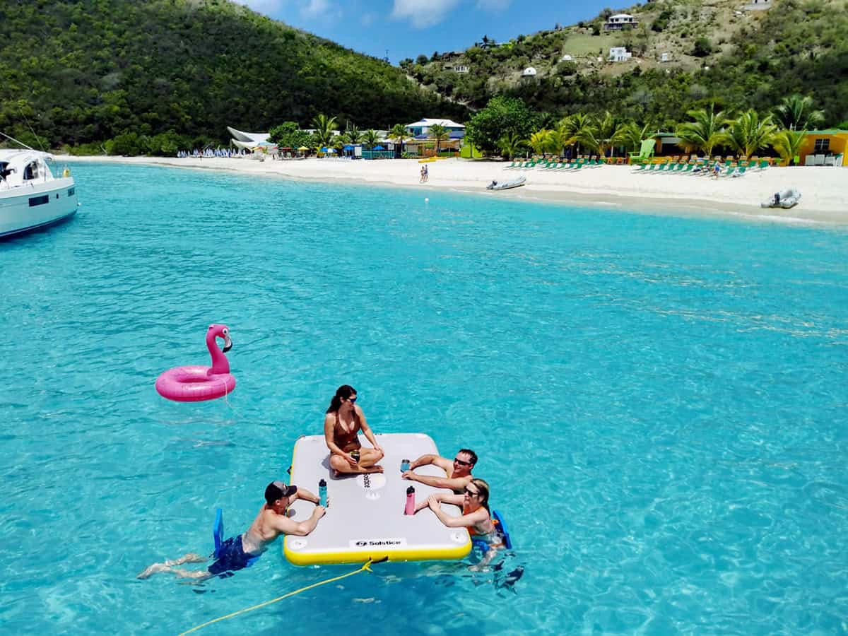A floating mat with 4 people enjoying drinks in bright turquoise water with white sand beach in background in White Bay, Jost Van Dyke, BVI