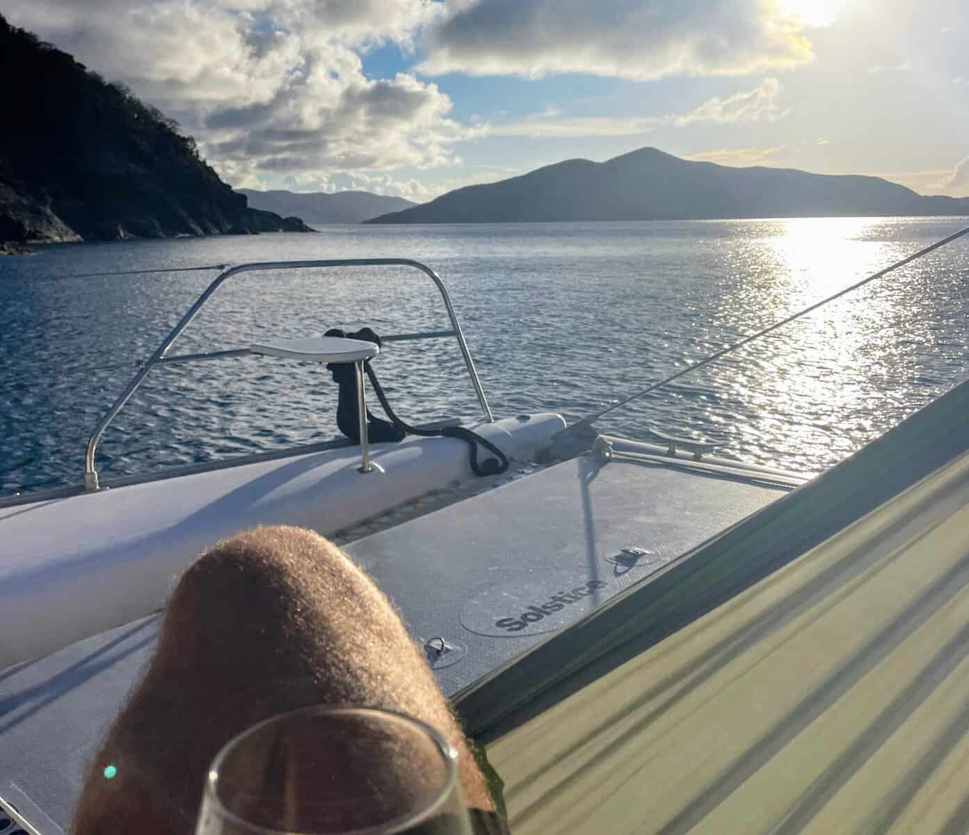 Person on a hammock on a sailing yacht with man's knee and wine glass and view of the sun shining on the ocean in the British Virgin Islands