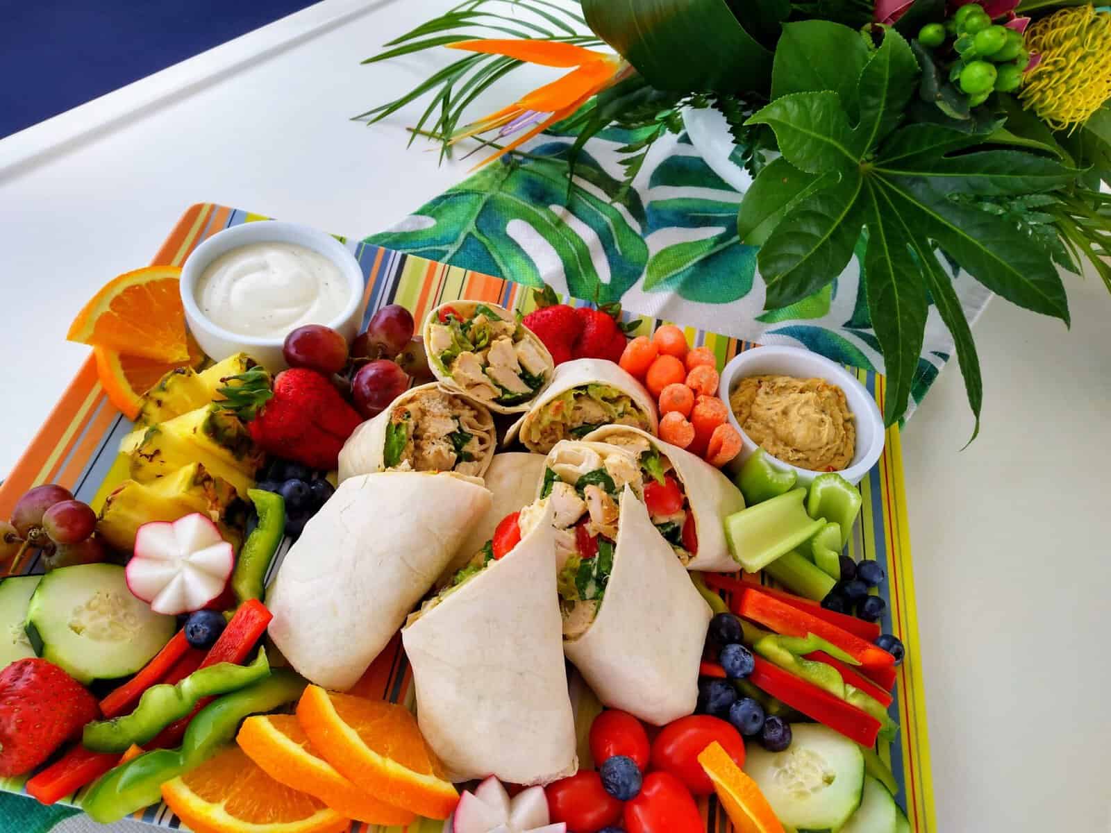 Brightly colored provisions tray with fruit, veggies, and chicken wraps on a yacht table with flowers