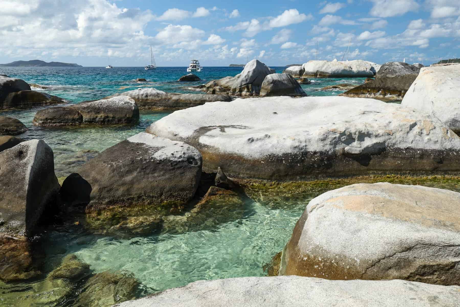Boulders and teal water with sailboats in the background at The Baths in BVI