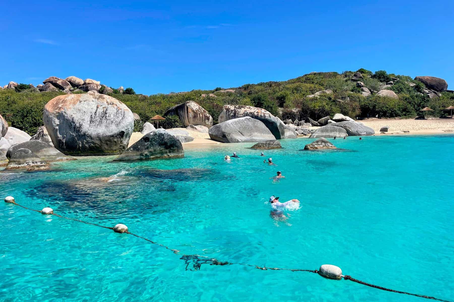 People swimming in bright turquoise water into a beach with boulders at The Bath in BVI