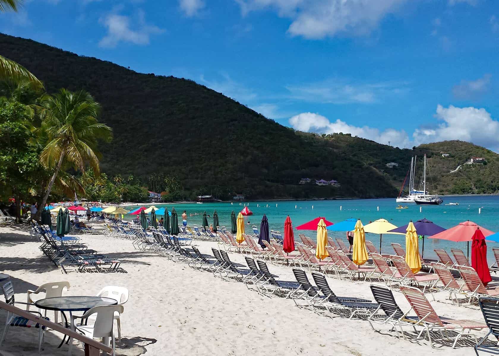 Cane Garden Bay, Tortola, British Virgin Islands. Colorful beach umbrellas with lounge chairs along teal ocean and green hillside.