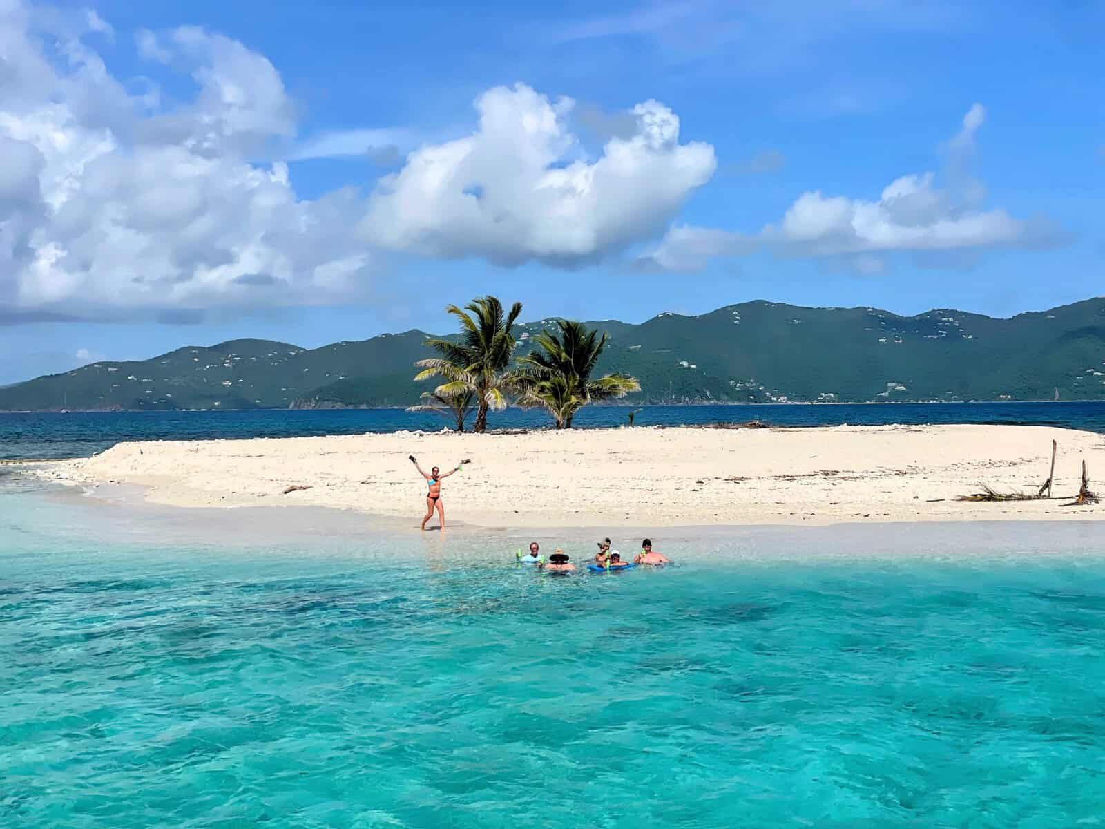 People playing in the ocean at Sandy Spit, British Virgin Islands. Sandy beach with two palm trees and teal water with green hillside of Tortola in the background