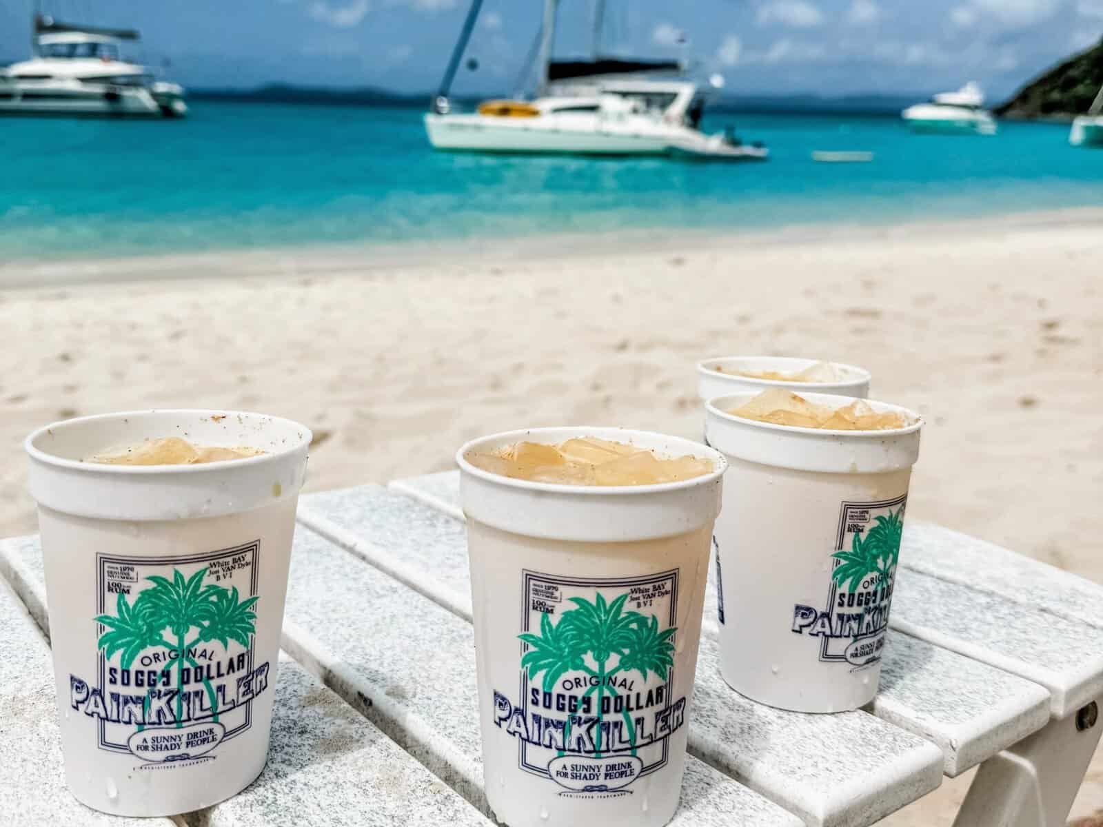 Four painkiller cocktails in Soggy Dollar cups with blue ocean and sailboats in background