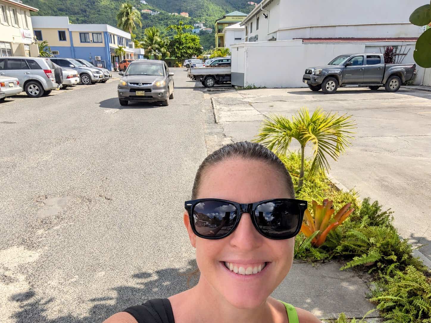 Mandy walking on a back street in Road Town, Tortola alone during the day with cars, buildings, and palm trees around. 