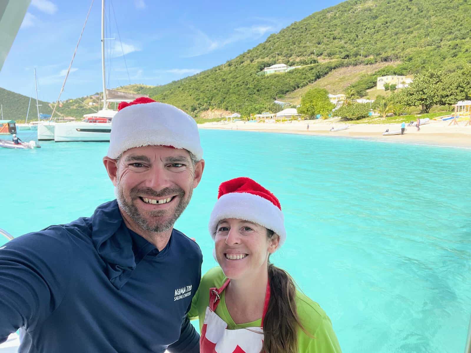 Joe and Mandy wearing Santa Hats in December in the Caribbean with bright teal water and white-sandy beach in the background
